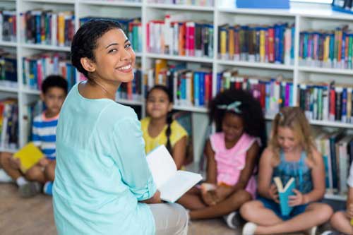 Teacher holding a book smiling in front of a classroom of students. 